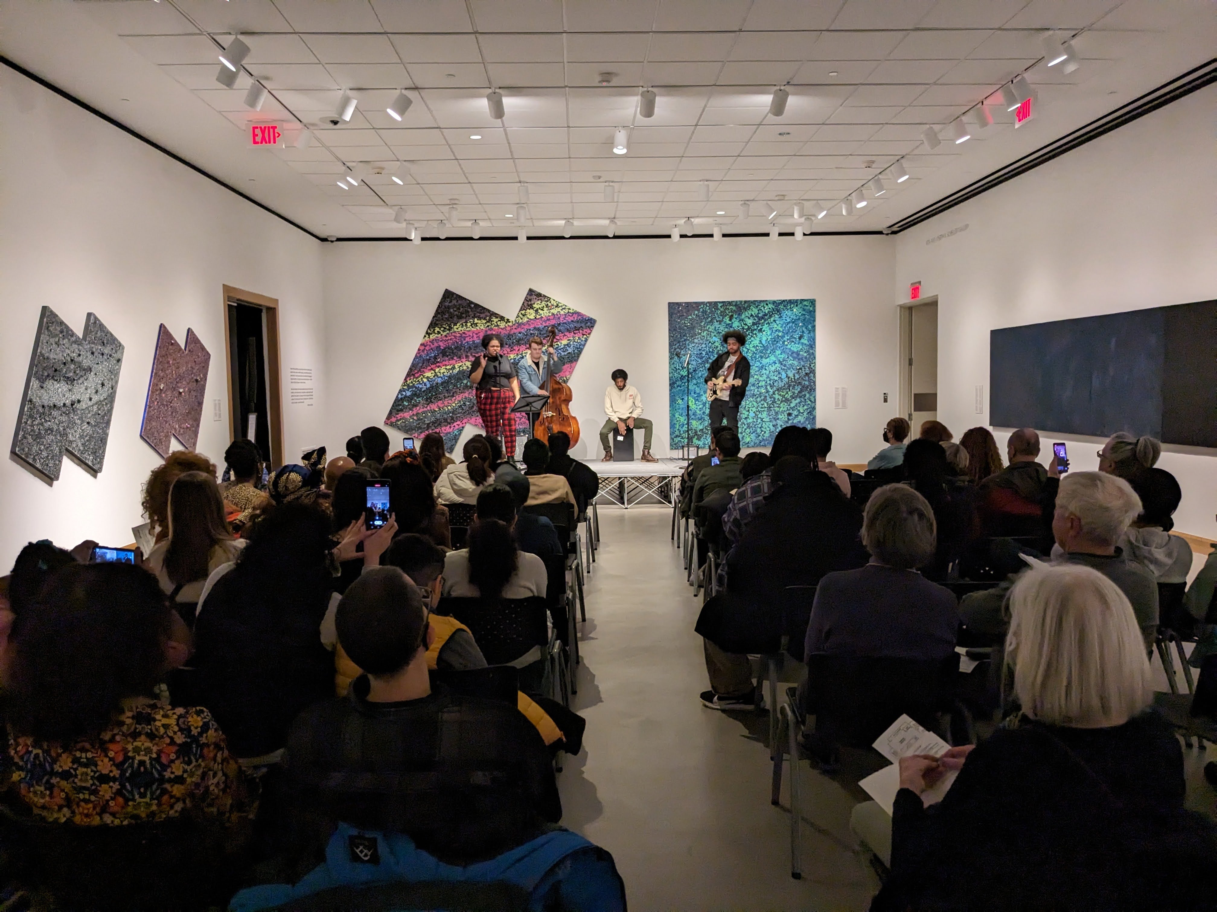 Four people, three playing instruments and one singing, stand on a stage in front of an audience. On all of the walls are large abstract paintings, each focusing on one or two diff...